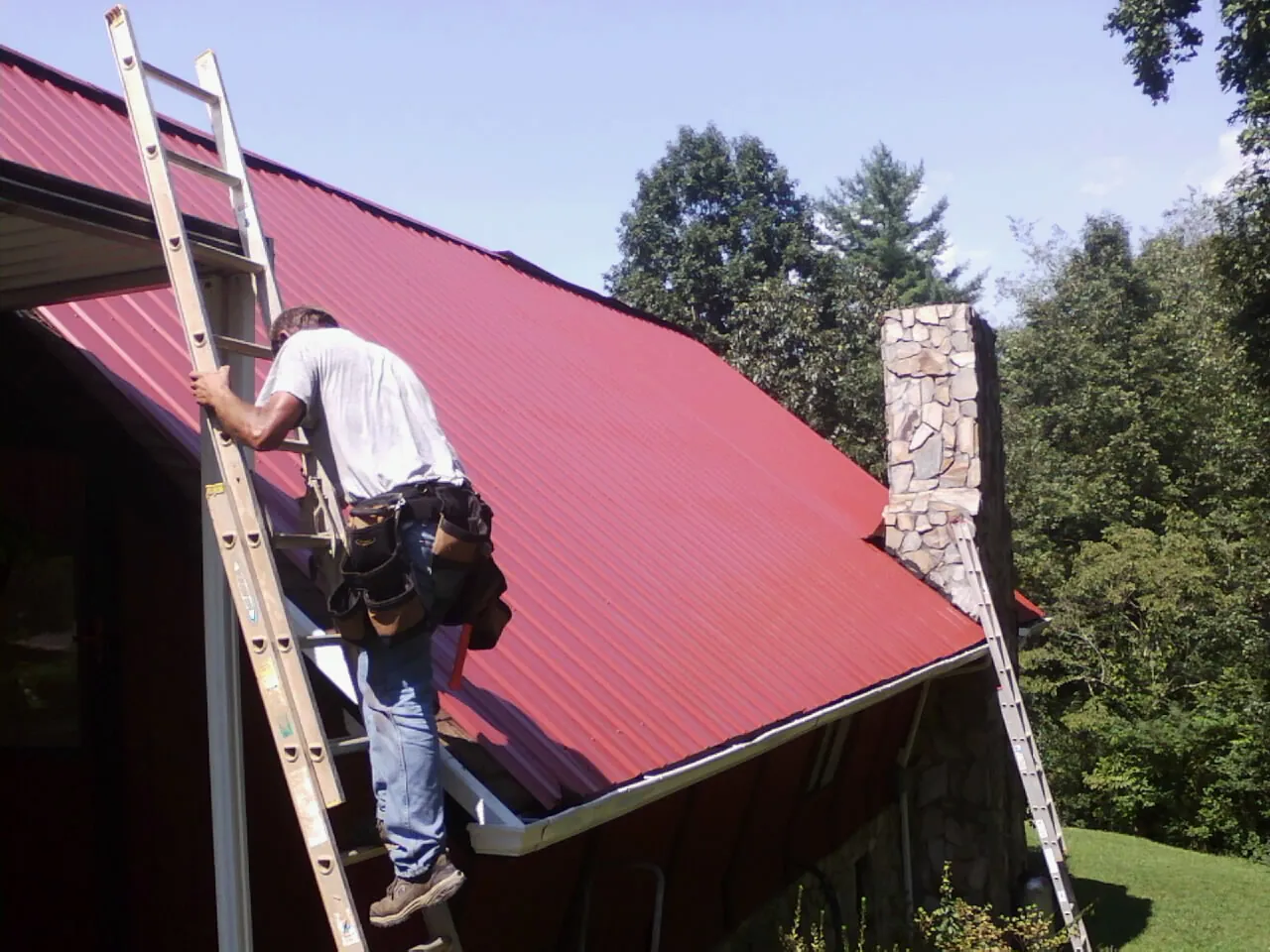 Roofer on a ladder beside a red metal roof and stone chimney — Soffit & Fascia Repair by Roofing Brothers in Kennedy